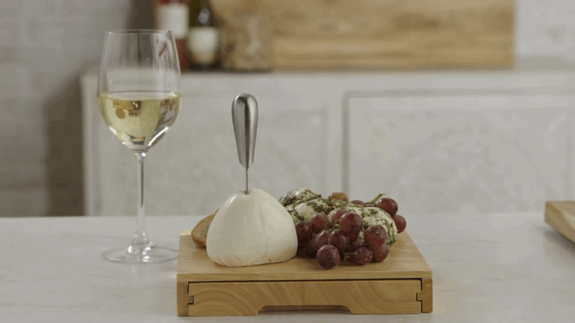 A glass of white wine sits next to a cheese ball, grapes, and herb-covered mozzarella on a wooden board